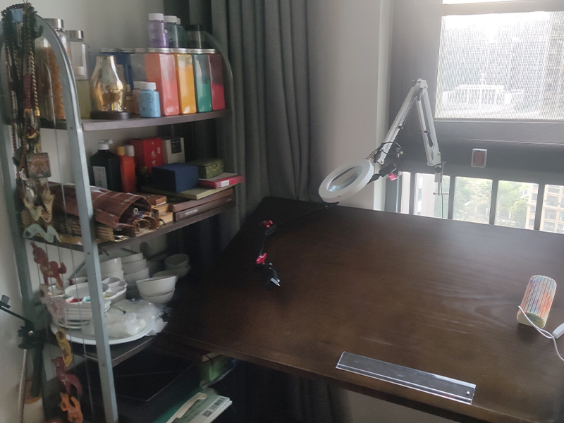 A serene and organized artist's workspace with a wooden desk, magnifying lamp, shelves of colors, and natural light from a window.