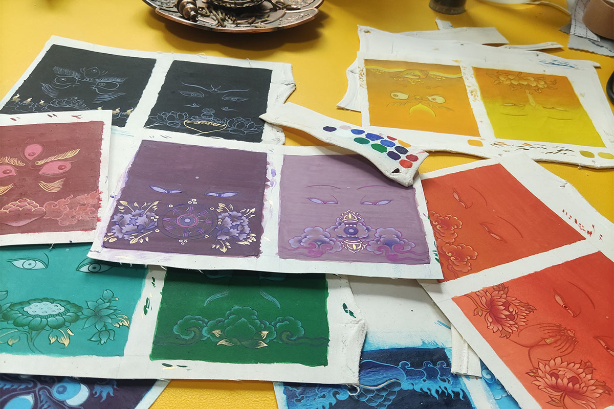 A close-up tabletop view of multiple hand-painted pure color Thangka cards from the Buddha Eyes series, overlapping each other alongside an artist color testing strip during the creative process.
