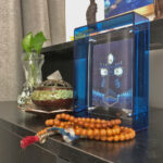Black Jambhala Buddha Eyes Thangka in a blue translucent acrylic frame styled on a desk with prayer beads.