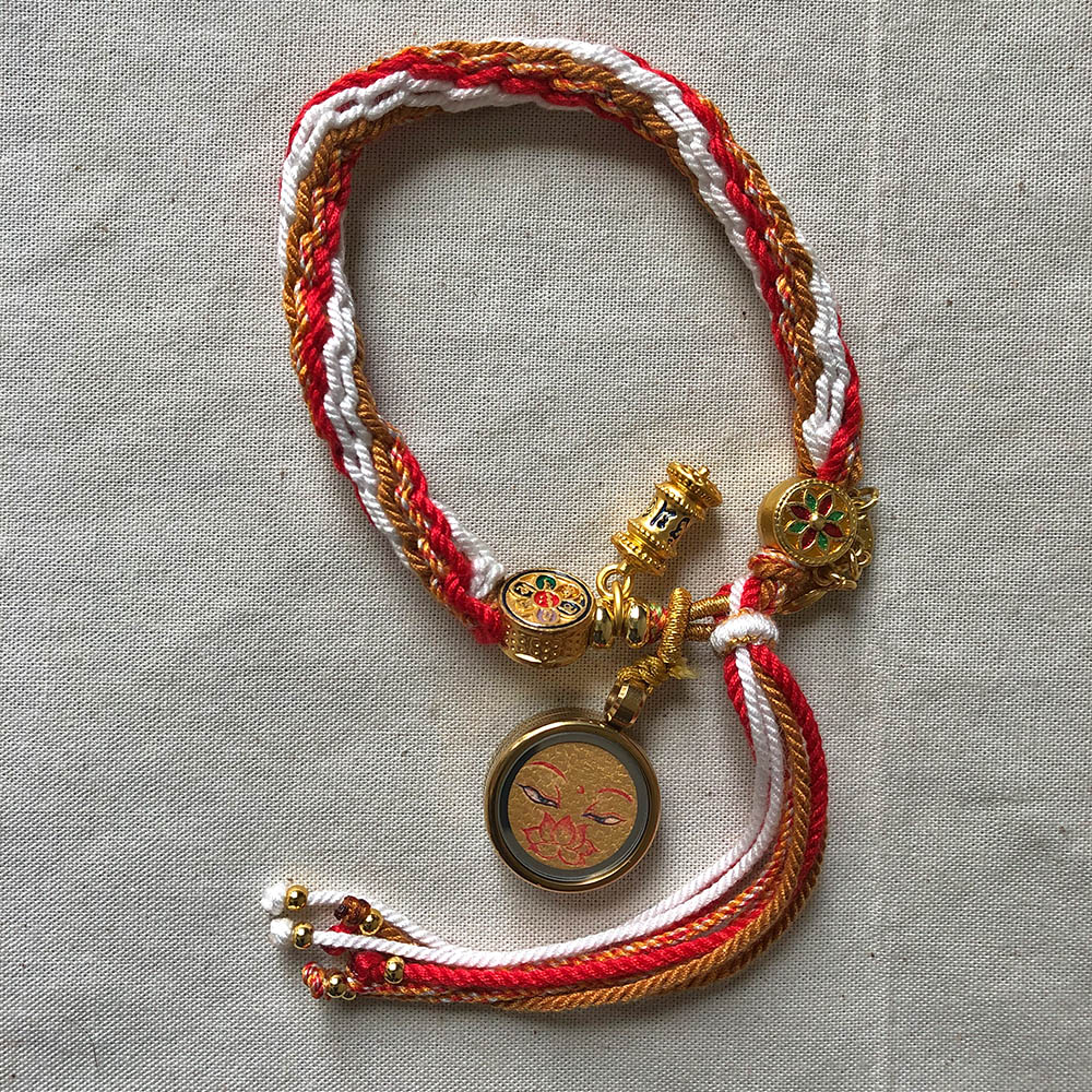golden-shakyamuni-buddha-eyes-orange-braided-meditation-bracelet-flatlay Golden Shakyamuni Buddha Eyes Tibetan braided meditation bracelet flatlay on fabric.