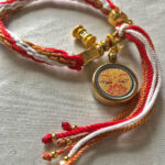 Angled view of Golden Shakyamuni Buddha Eyes Ghau pendant attached to red and white braided cord.