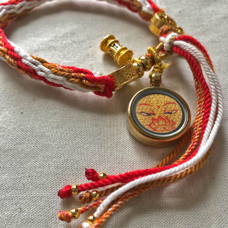 Angled view of Golden Shakyamuni Buddha Eyes Ghau pendant attached to red and white braided cord.