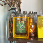 Green Jambhala Thangka in a yellow translucent acrylic frame styled on a desk with ambient lighting.