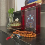 Indigo Yamantaka Buddha Eyes Thangka in a red translucent acrylic frame styled on a desk with prayer beads.