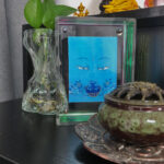 Medicine Buddha Eyes Thangka in an iridescent translucent acrylic frame styled on a desk with an incense burner.