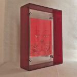 Side perspective of orange Avalokiteshvara Buddha Eyes art inside a red translucent acrylic frame.