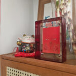 Orange Avalokiteshvara Buddha Eyes Thangka in a red translucent acrylic frame styled on a desk with fortune cat.