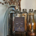 Padmasambhava Buddha Eyes Thangka in a black translucent acrylic frame styled on a shelf with glass bottles.