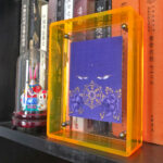 Purple Vairochana Buddha Eyes Thangka in a yellow translucent acrylic frame styled on a bookshelf.