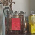 Red Amitayus Buddha Eyes Thangka in a red translucent acrylic frame styled on a shelf with glass bottles.
