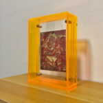 Red Jambhala Thangka print in an amber yellow translucent acrylic stand on a wooden table.