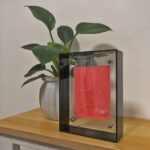 Shakyamuni Buddha Eyes Thangka in a black translucent acrylic frame styled on a wooden desk with a plant.