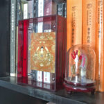 Shakyamuni Buddha Thangka in a red translucent acrylic frame styled on a bookshelf next to books.