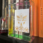 White Jambhala Buddha Eyes Thangka in an iridescent acrylic frame styled on a bookshelf next to books.