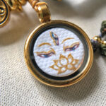 Close-up of White Tara Buddha Eyes pendant with hand-drawn gold linework and lotus charm.