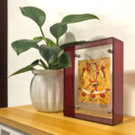 Yellow Jambhala Thangka in a red translucent acrylic frame styled on a desk with a green plant.