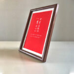 Side perspective view of A4 dark wood framed red calligraphy print on tabletop