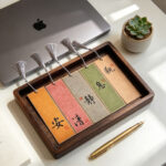 Set of 5 colorful handwritten Chinese calligraphy bookmarks neatly arranged inside a dark wooden tray near a brass pen and laptop.