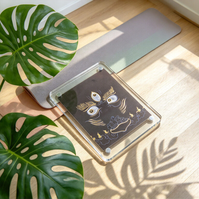 A black Buddha Eyes acrylic art card flat lay surrounded by tropical monstera leaf shadows in natural sunlight.
