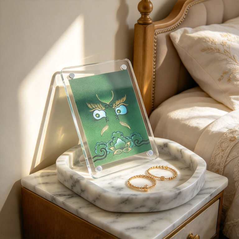 A Green and gold Buddha Eyes acrylic art card standing on a marble tray near a bedside lamp.
