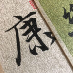 Extreme macro close-up of handwritten Chinese character Kang Health with red artist seal on grey Xuan paper card.
