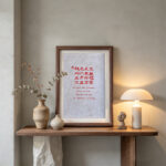 Framed calligraphy artwork A Home in Harmony on a dark wooden hallway shelf beside a modern lamp and dried flowers.