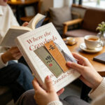 Hands holding an open novel with two calligraphy bookmarks tucked between pages, showing handwritten Asian characters.