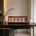 Horizontal dark wood Song Brocade calligraphy frame on a modern black console table with greenery and twin candles.