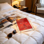A red Buddha Eyes acrylic pocket art card standing upright on a white duvet in a warm sunlit bedroom.