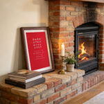 Dark wood framed red romantic calligraphy print on a table beside a burning fireplace and a lit candle.