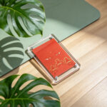 A orange Buddha Eyes acrylic art card laying flat on a wooden surface next to a green yoga mat and tropical palm shadows.